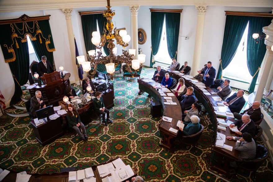 In a green patterned carpeted room are curved wooden desks full of people, as seen from above