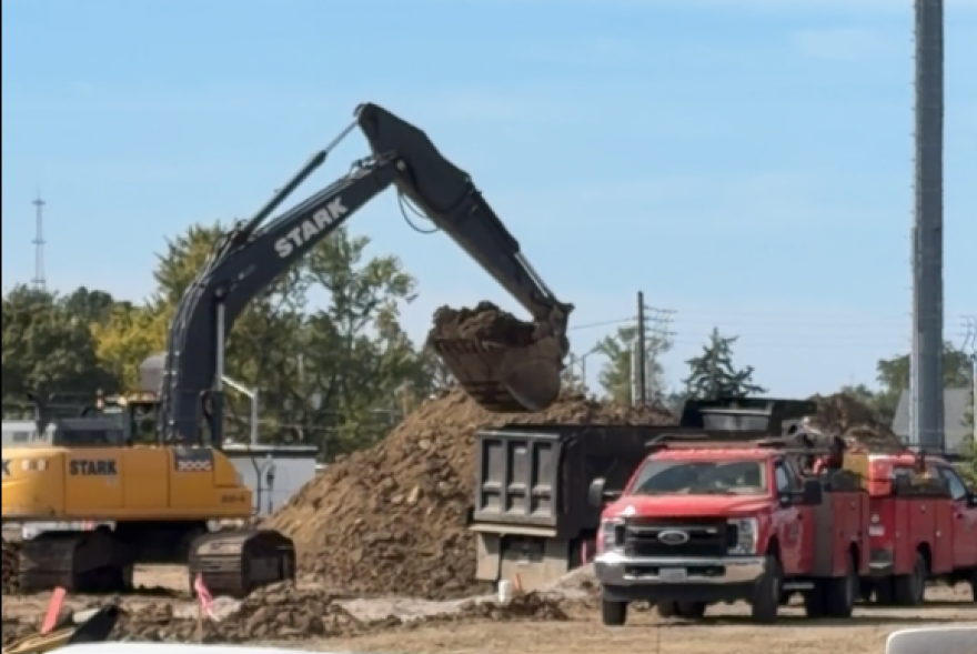 a yellow backhoe dumps dirt into a truck parked next to red pickup trucks