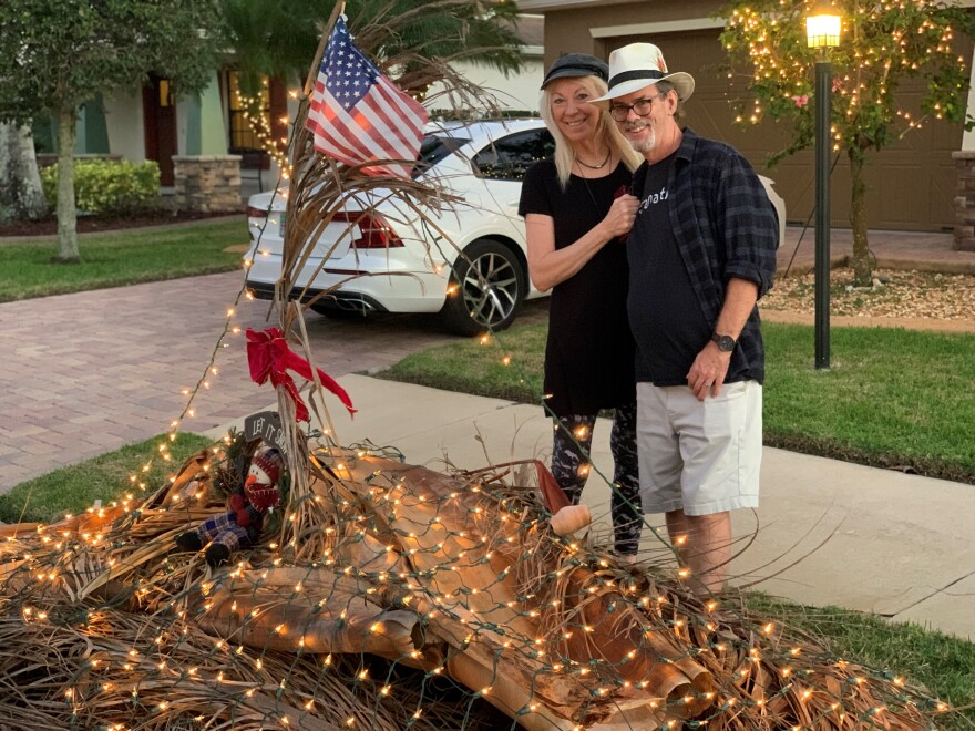 Kathy and David Thomas pose by their decorated debris pile on Friday, December 2, 2022. It has white lights, a wreath and a red bow, and an American flag on it.