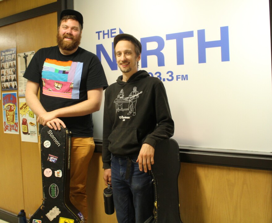 Two smiling men stand in front of a frosted window that says the North 103.3 FM in blue lettering. They are both leaning on guitar cases.