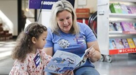 A blonde mid-aged woman, presumably a Veterans United employee, reads a picture book to a young girl with curly brown pigtails and a pink patterned dress. 