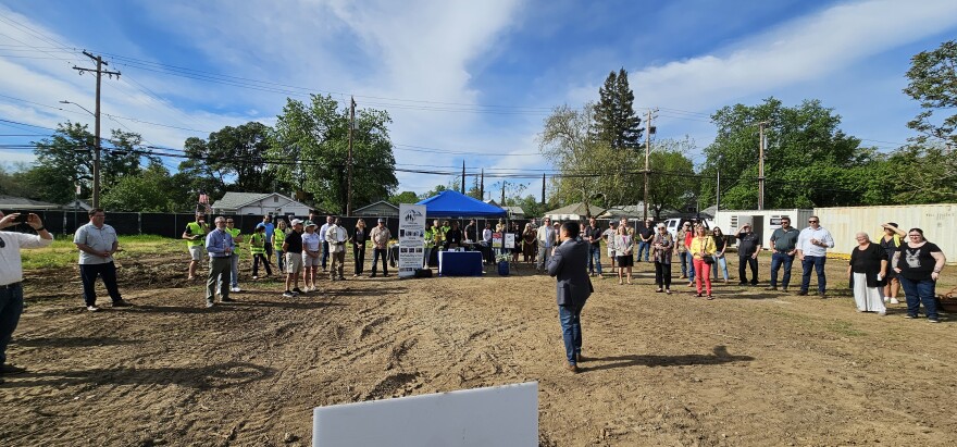 A crowd of people are gathered on a dirt lot while a man speaks into a microphone under a blue sky. 
