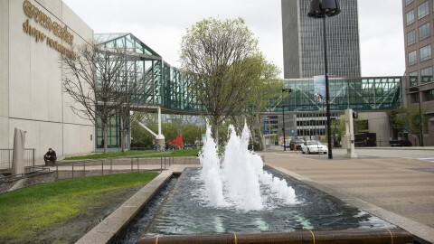 An urban location shows a grassy field in the foreground with many tall buildings and a park in the background.