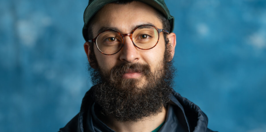Close-up photo of Ameen Wahba. He's wearing glasses, a baseball hat and has a beard. He's standing against a blue backdrop.