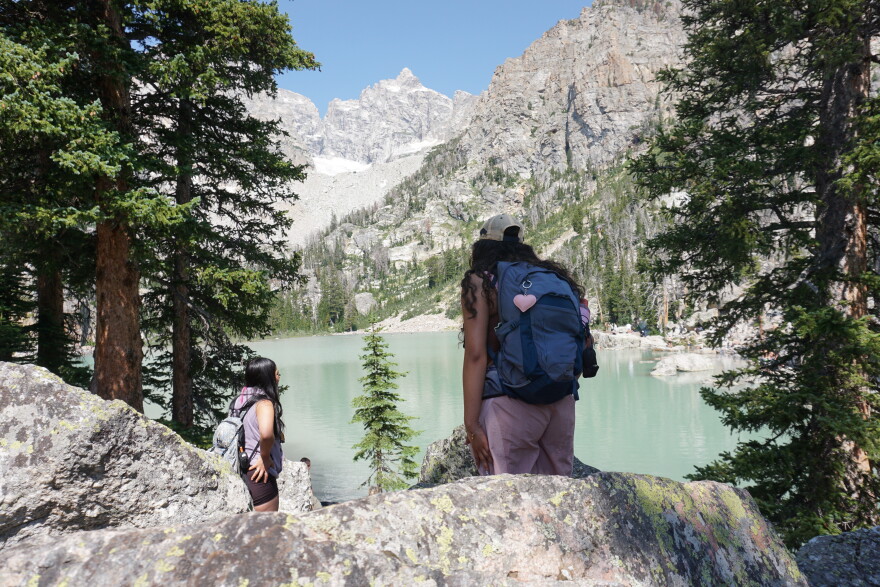 Two women look towards an aquamarine colored lake with mountains above.