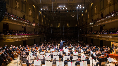 A view from the orchestra, looking over a sea of sheet music and stands. Andris Nelsons stands on a podium with his arms in the air. Behind him, a see of audience members and the vastness of Symphony Hall.
