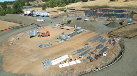 An overhead view of a dirt construction site is shown, with piles of materials as well as construction vehicles. 