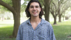 man with long hair and glasses stands in front of trees
