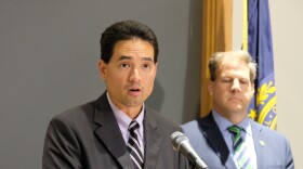 A man speaks at a microphone, with another man standing in the background. 