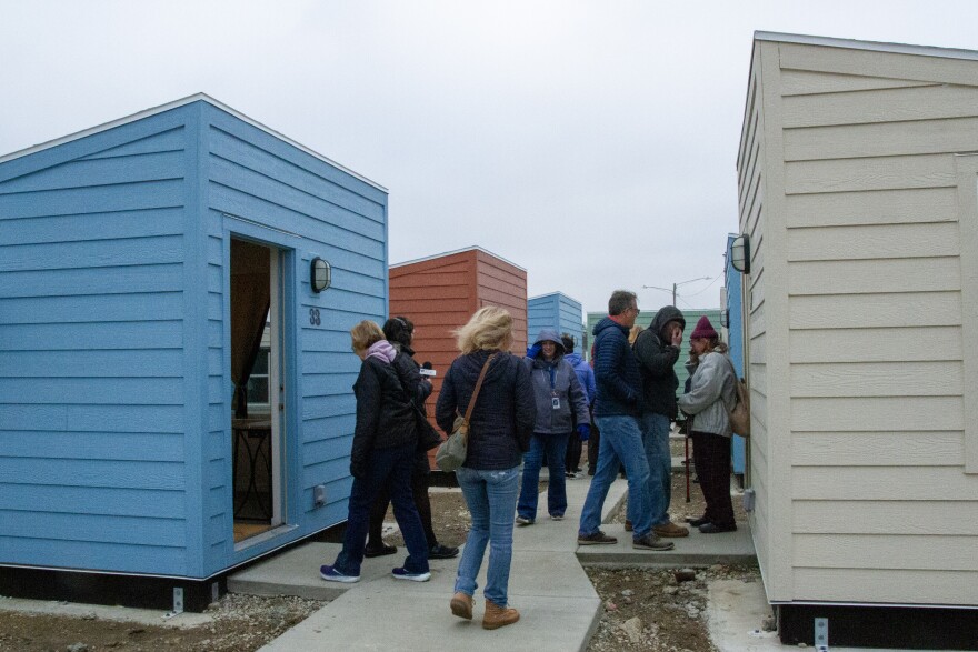 On a cloudy day, a group of people tours colorful small cabins separated by new concrete sidewalk