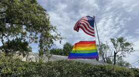 Rainbow LGBTQ pride flag and American flag wave on a pole above a home surrounded by tRainbow LGBTQ pride flag and American flag wave on a pole above a home surrounded by trees