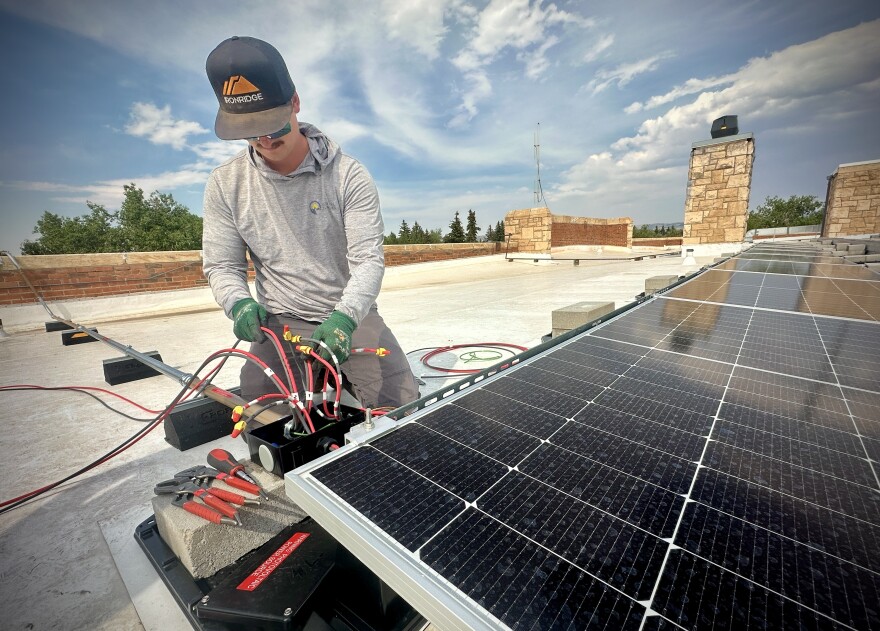 A man in a grey shirt on a roof next to a black solar panel and red electrical chords.
