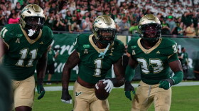 3 football players wearing green jerseys and gold helmets and pants running together, with player in middle carrying football in left hand