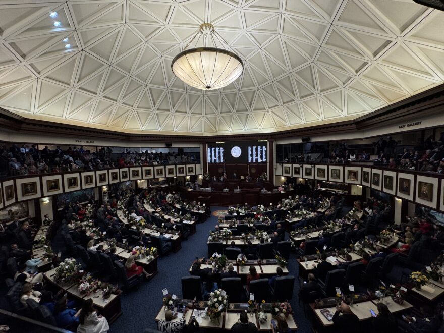 Lawmakers gathered in the Florida House of Representative chambers. A desk in the middle as other tables circle it in a round room with a dome ceiling. blue carpeting, white and brown paneling