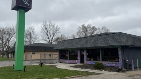 On a cloudy day, a building with a purple swirly paint pattern and a black shingled roof can be seen. In front of the building, a large green sign reads "Doja".