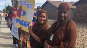 Two Somali women with brown head coverings in the forefront of a photo of people lining a sidewalk. One woman is holding a sign that reads "Thank you law enforcement."