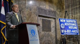 A man in a grey suit with a gold tie stands behind a wooden podum with a opaque sign on it that has the seal of Jackson County. In the right of the photo is a blue sign with details about a tax assistance hotline.