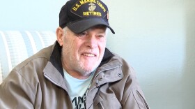 A man, Jerry Hart, sits in an arm chair. He has a hat on that says "US Marine Corps Retired"