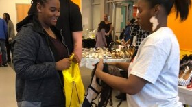 Two Black women facing each other.  The woman on the left is holding open a yellow drawstring bag as the woman on the right gets ready to put several bras in the bag.
