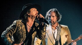 A man in a paisley-print jacket and black wide-brimmed hat sings into a microphone on stage while another man in a gold jacket plays electric guitar next to him.