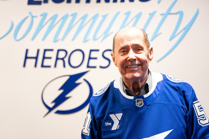 Older bald man wearing blue hockey jersey and smiling into the camera with a logo of a lightning bolt behind his right shoulder