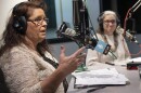 A woman seated left, gestures with her right hand while talking at a microphone studio while another woman listens.