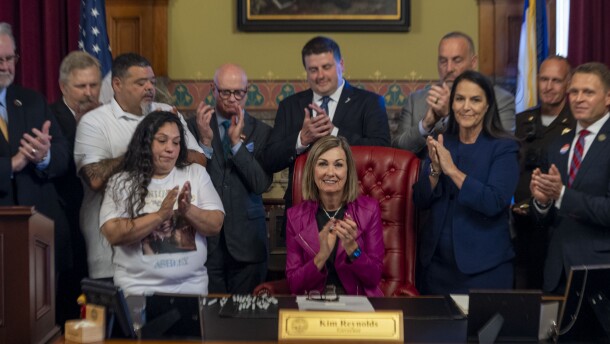 a woman sits at a desk and several other people stand around her as they all clap