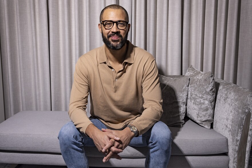 a man sits on a gray couch wearing jeans, a camel-colored long-sleeve polo shirt and glasses