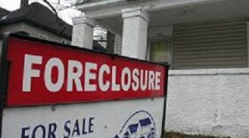 A photograph of an old white house with sign out front reading "Foreclosure" and "For Sale."