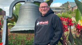 A man in a black "toys for tots" sweatshirt stands in front of a bell and flower beds, smiling at the camera.