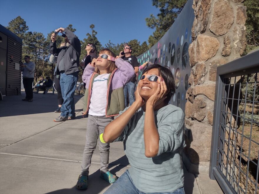 A woman and young girl look skyward, wearing eclipse glasses. 