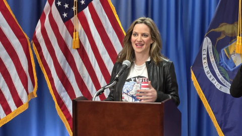Michigan Secretary of State Jocelyn Benson, in a white shirt and black jacket, speaks at a lectern bearing the state of Michigan seal.