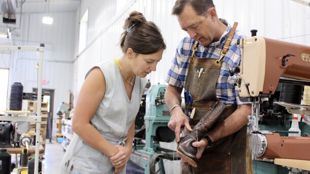 A man in a buttoned-down shirt and apron holds up a cowboy boot being stitched together to show a woman in a denim jumper.