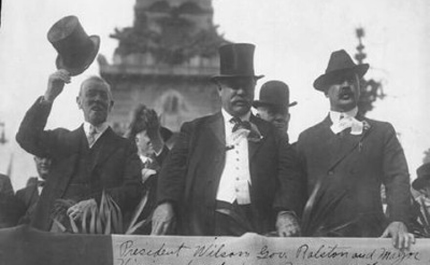 Historical black and white photograph from 1916 showing President Woodrow Wilson tipping his top hat alongside Indiana Governor Samuel M. Ralston and Indianapolis Mayor Joseph E. Bell as they view a parade from a reviewing stand in front of the Soldiers and Sailors Monument.