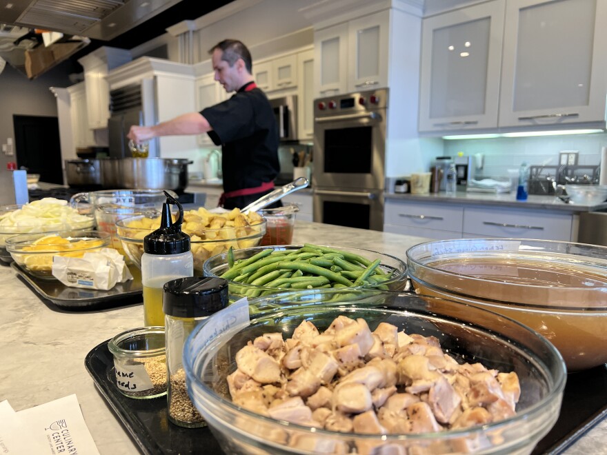 Instructor and manager at the Dorothy Lane Market Culinary Center, Ryan Noreikas, cooks in the background with trays of pre-portioned food in the forefront