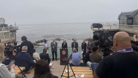 Six nicely-dressed dignitaries stand along a glass fence with Monterey Bay churning behind them. In the foreground, on a slope with bench seats, a handful of people sit with notebooks and cameras. 