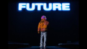 A man wearing winter clothing stands on stage with his back to the camera in front of a large screen that reads "FUTURE."