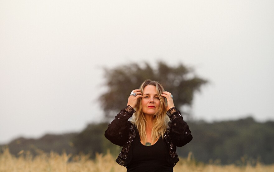 A woman poses in a field with her hands up by her head. She has dirty blonde hair and black clothes on.
