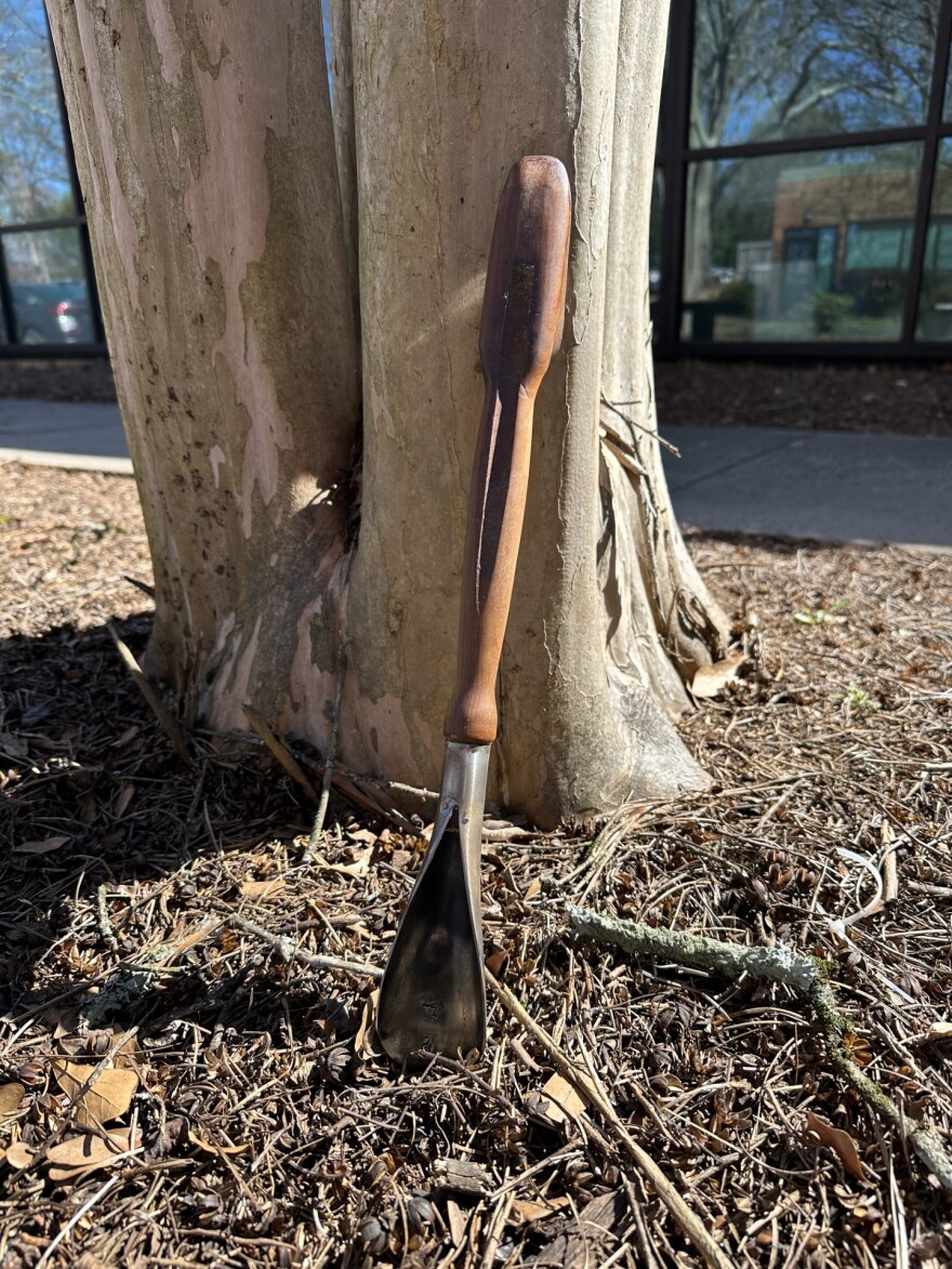 A handmade garden trowel with a wooden handle stuck in the soil at the base of a tree.