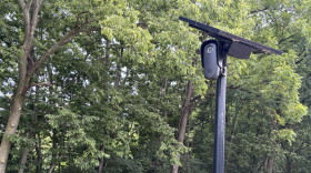 A black license plate reader and solar panel sit on the top of a pole, with woods in the background.