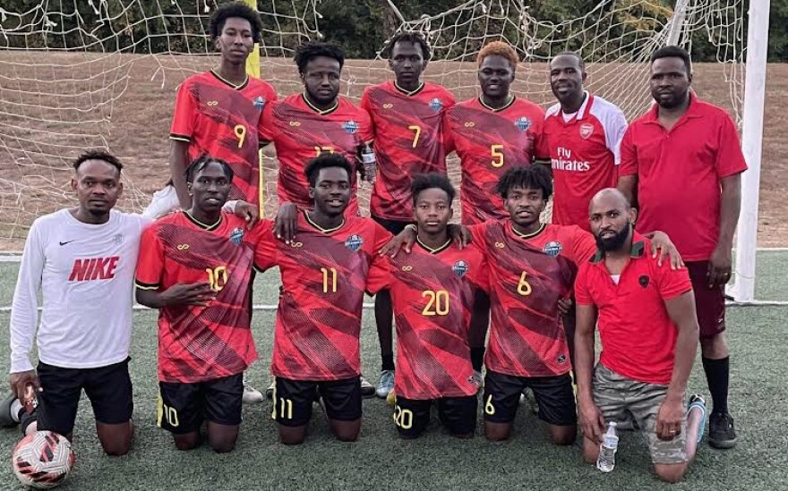 Twelve men, six kneeling and six standing, pose for a group photo near a soccer goal. They are wearing red, patterned jerseys with yellow numbers and black shorts.