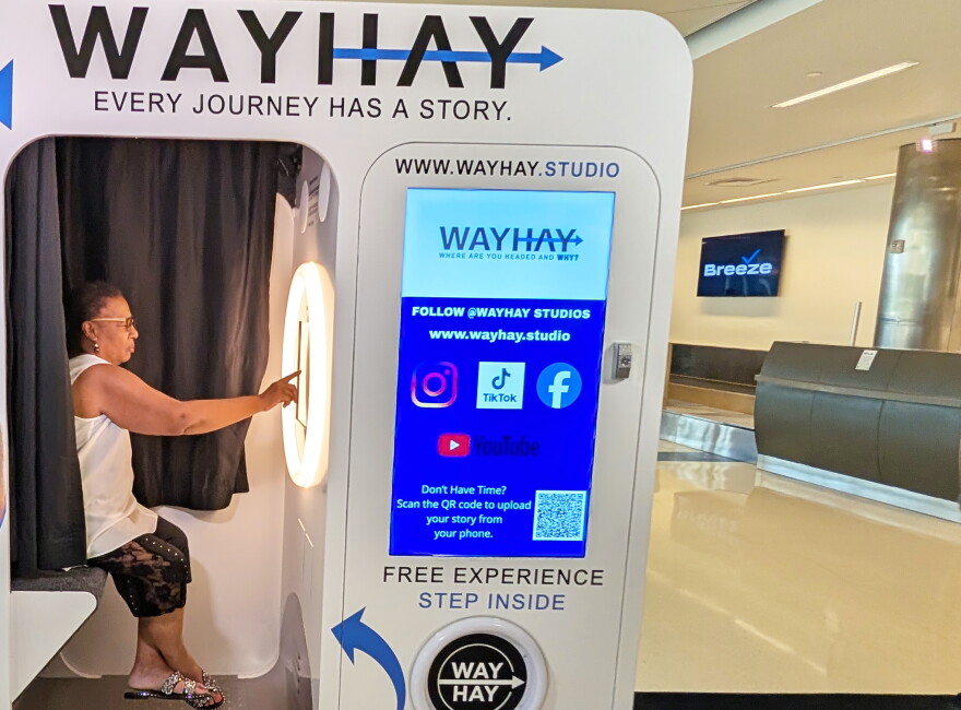 A woman sits inside a video booth and reaches out with one arm to tap the screen in front of her. The booth is decorated with blue and black arrows and text against a white background. "WAYHAY" and the tagline "Every Journey Has A Story" are at the top of the booth, over the doorway to the compartment where the woman is sitting.