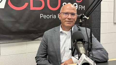 A man in a gray suit and glasses sits at a radio station desk with a microphone. The background shows a WCBU 89.9 FM banner and part of a white brick wall.