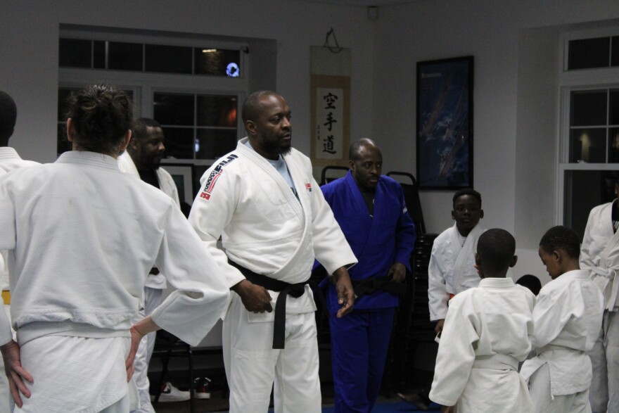 A judo instructor wearing a white uniform and a black belt looks out over a group of judo students. 