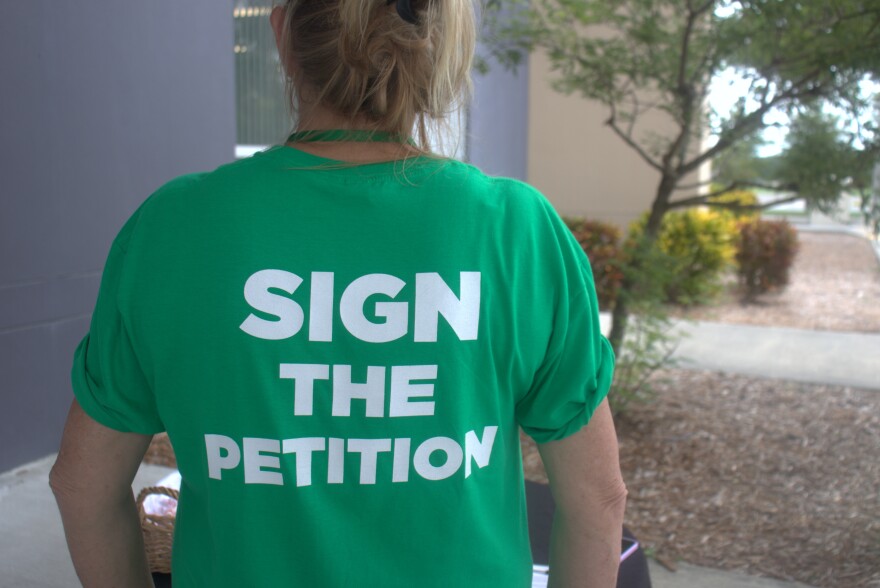 A woman, standing in front of tree, wears a green T-shirt that says "SIGN THE PETITION." She's only visible from behind.