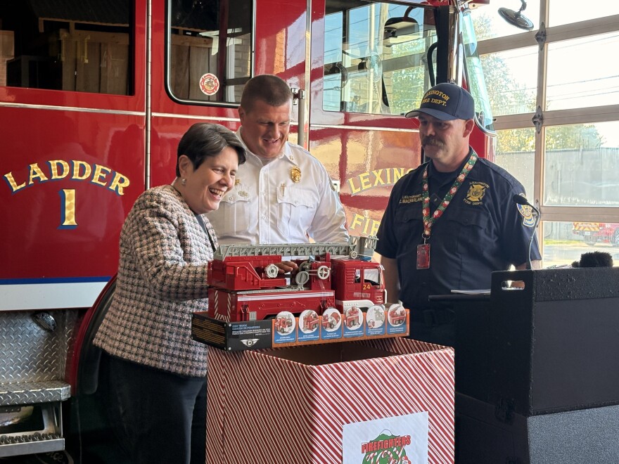 Mayor Linda Gorton dropped off the first donation of this year's drive, demonstrating the horn and sirens of a toy fire truck to Assistant Chief Joey Harris and Lt. Chris MacFarlane. Gorton and Harris have wide smiles, while MacFarlane looks attentively cheerful. They stand behind a lectern and in front of a real-life fire truck in the station garage.