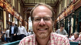 A bearded man with red-ish brown hair wears glasses and a red plaid shirt 