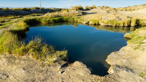 A small, round hot spring pool with dark blue water, surrounded by sandy soil, rocks, and desert grasses under a clear blue sky.