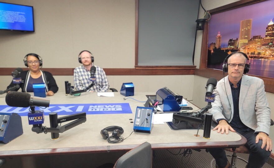 Three people wearing headphones sit at a table in a radio talk studio: A woman back left has her dark hair pulled back. She is wearing glasses and a dark top under a black sweater with a white border; a man back center has short red hair and is wearing glasses and a blue and white plaid button-down shirt; a man front right has very short dark hair and is wearing glasses, a black button-down shirt under a grey suitcoat, jeans, and brown shoes.