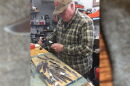 A man wearing a plaid patter shirt measuring silver knives with a table with a pile of knives in front of him.